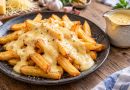 Goldene Pommes mit cremiger Käsesoße auf einem dunklen Teller, daneben eine kleine Kanne mit Käsesoße.